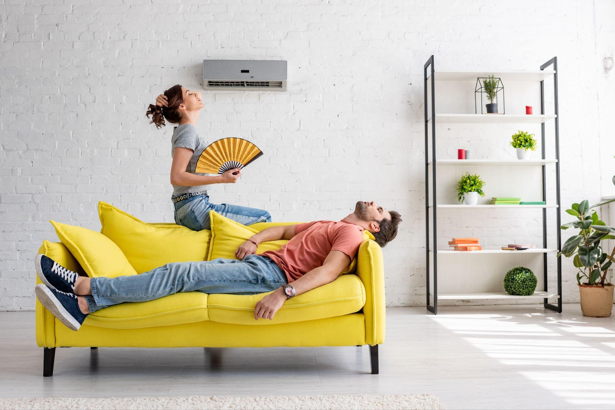 a man lying on yellow sofa and a lady sitting nearby on the sofa with a fan under an air conditioner